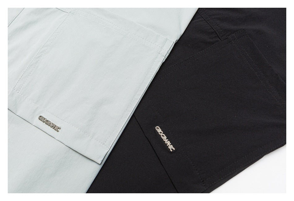 Close-up of pale gray-green and black shirt fabric showing stitched panels and small metal GEOGRAPHIC badges.