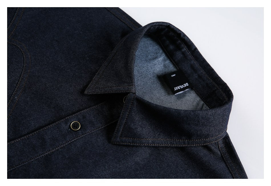 Close-up of a dark indigo denim collar and button with a black neck label, shown inside a white border.