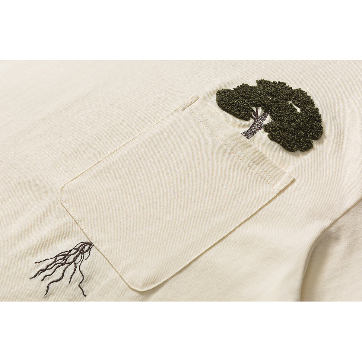 Close-up of an ivory T-shirt pocket with a dark embroidered tree canopy above it and roots below.
