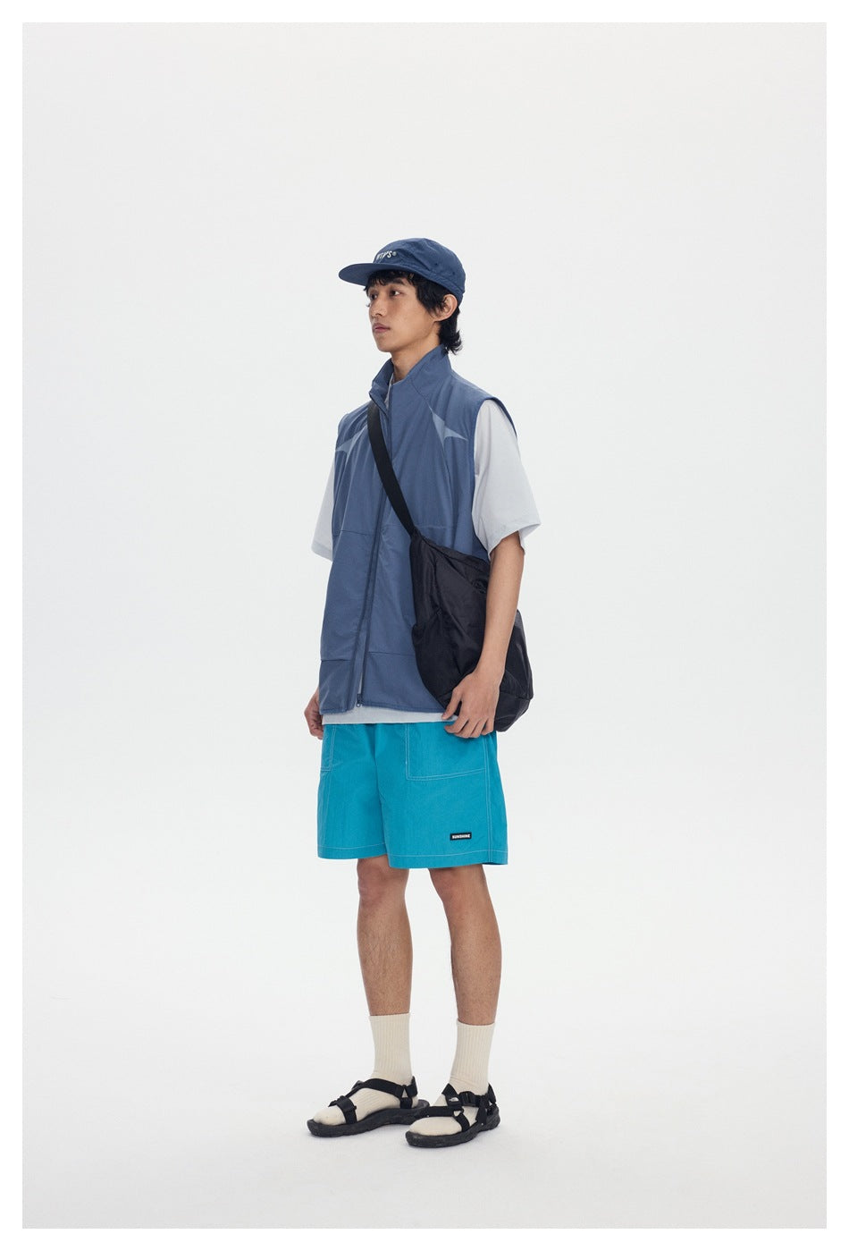 Model wearing a blue zip-up stand-collar vest over a light tee with aqua shorts, shown from the side