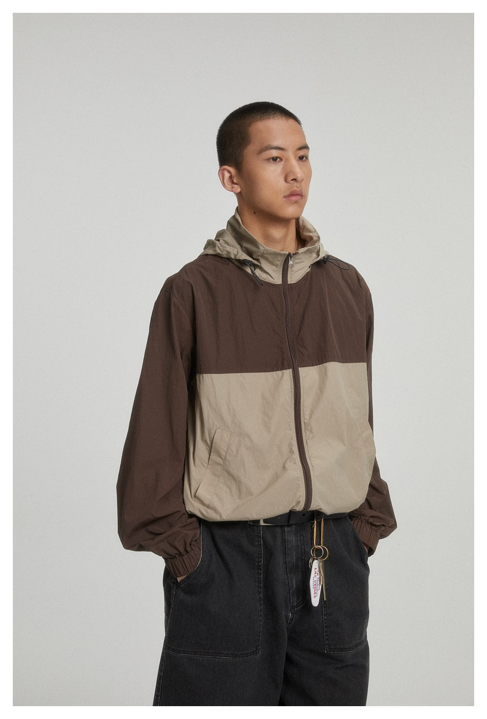 Model wearing a two-tone hooded jacket with a dark brown upper and sleeves, beige lower panel, and black trousers.