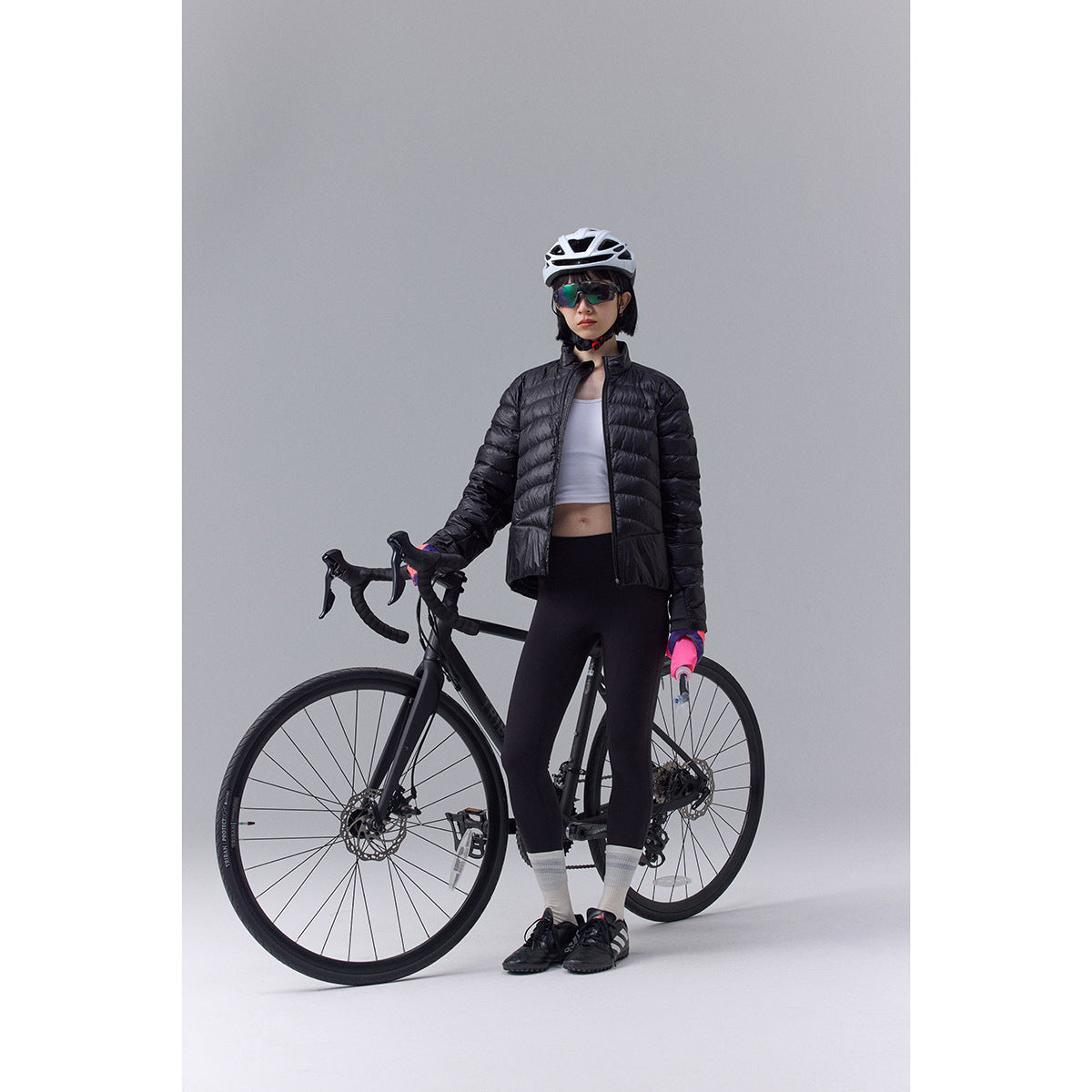 Model wearing a black quilted jacket and cycling gear beside a road bike against a gray studio background.