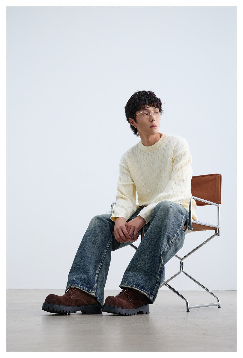 Seated model wearing an ivory cable-knit crewneck sweater and dark blue jeans.