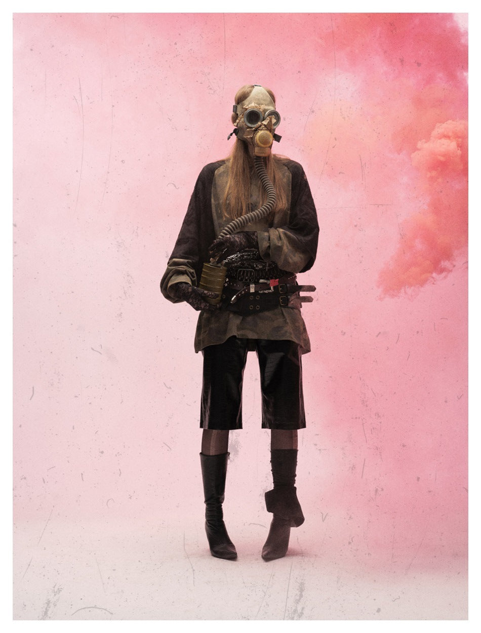 Model in a gas mask and belts wearing a camouflage top with dark sleeves and black shorts in pink smoke