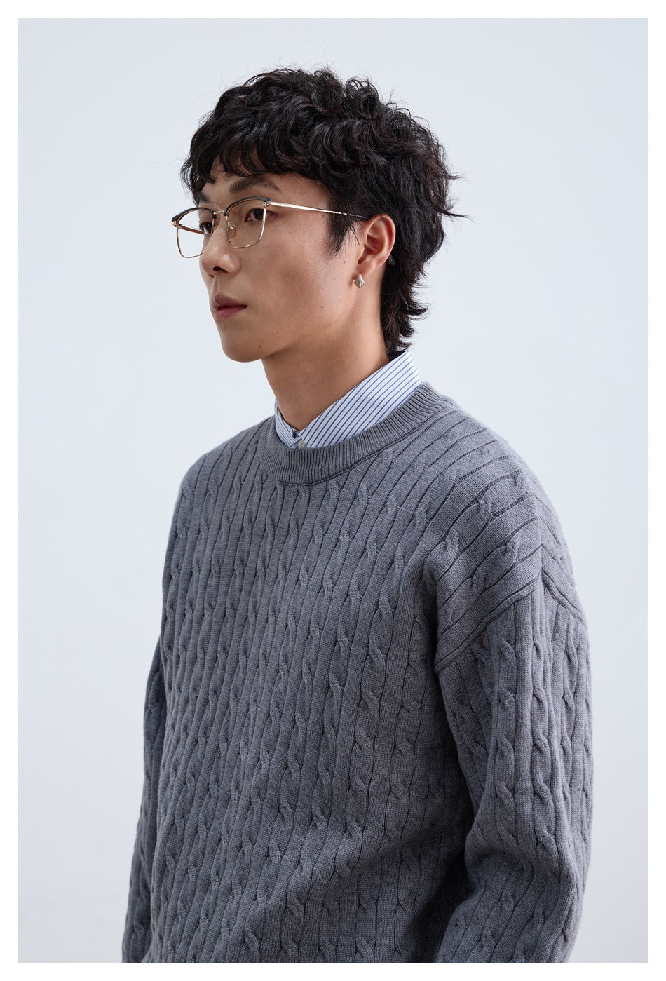 Close view of a model wearing a gray cable-knit crewneck sweater over a striped collared shirt.