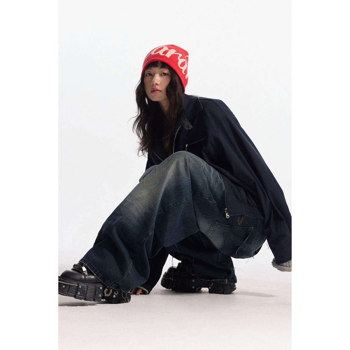 Model crouches in a dark navy zip jacket, oversized dark jeans, black boots, and a red knit beanie.