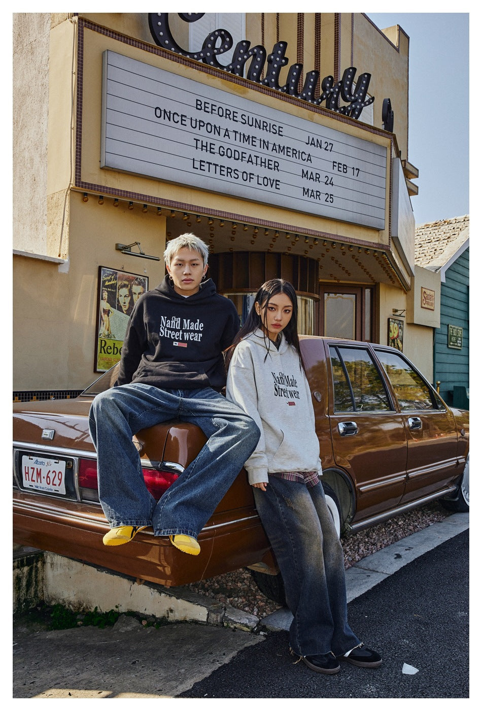 Two models sit by a car wearing black and light gray graphic hoodies with loose jeans.