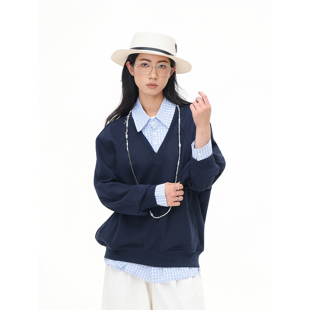 Model wearing a navy V-neck sweatshirt over a blue checked shirt with wide white trousers and a cream hat.