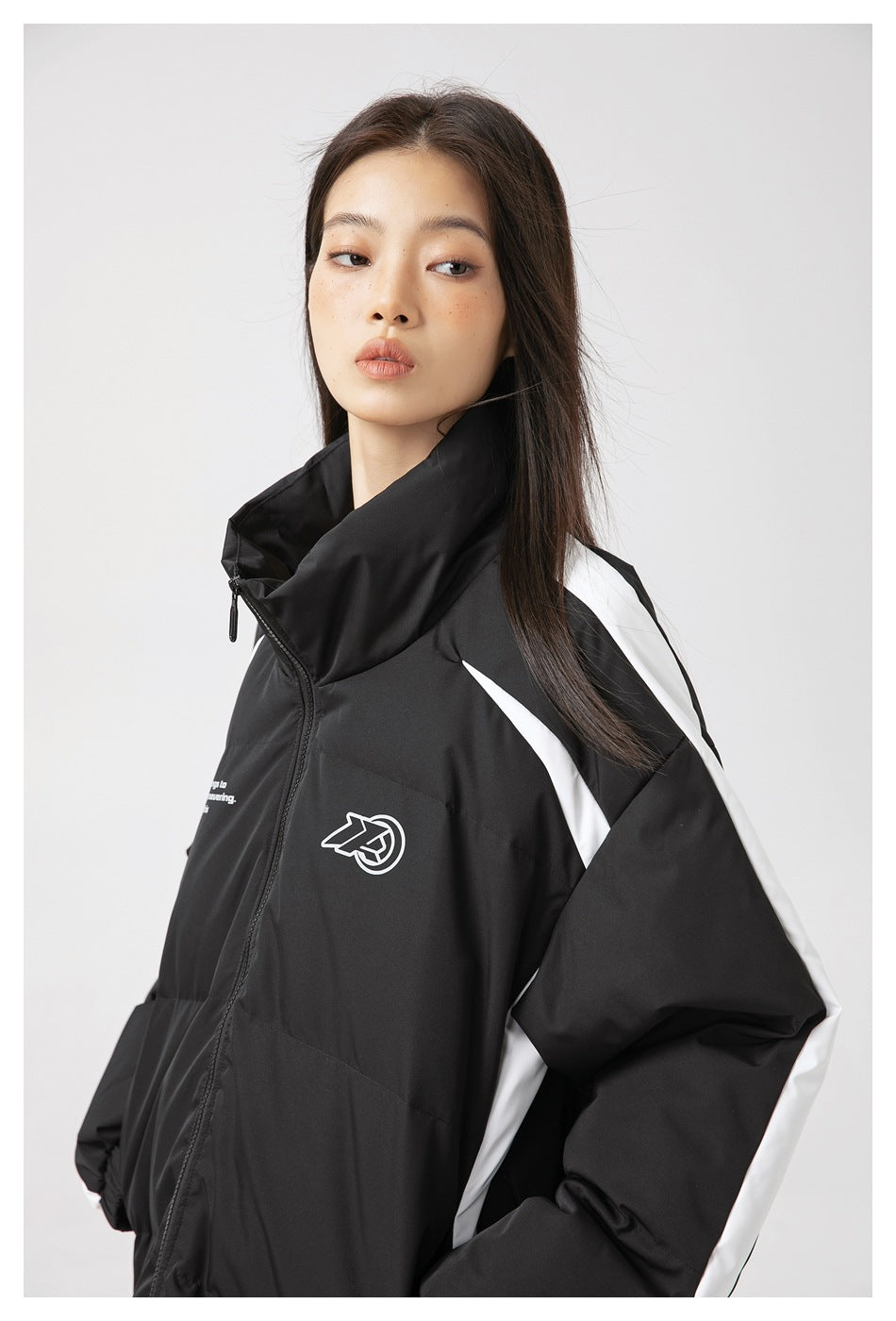 Three-quarter view of a model in a black padded jacket with a high collar, white sleeve stripes, and chest graphics.