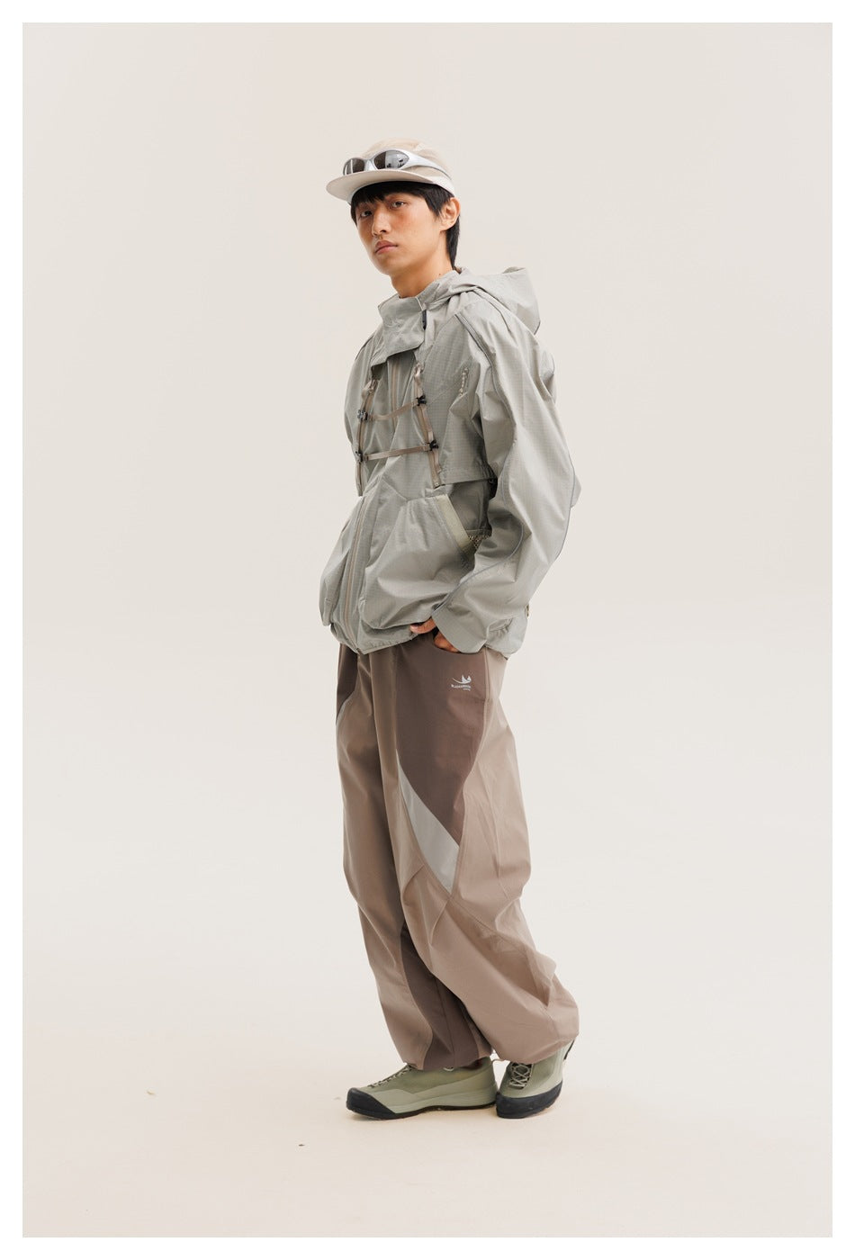 Side view of a model wearing a light gray hooded jacket with chest straps and two-tone taupe pants.