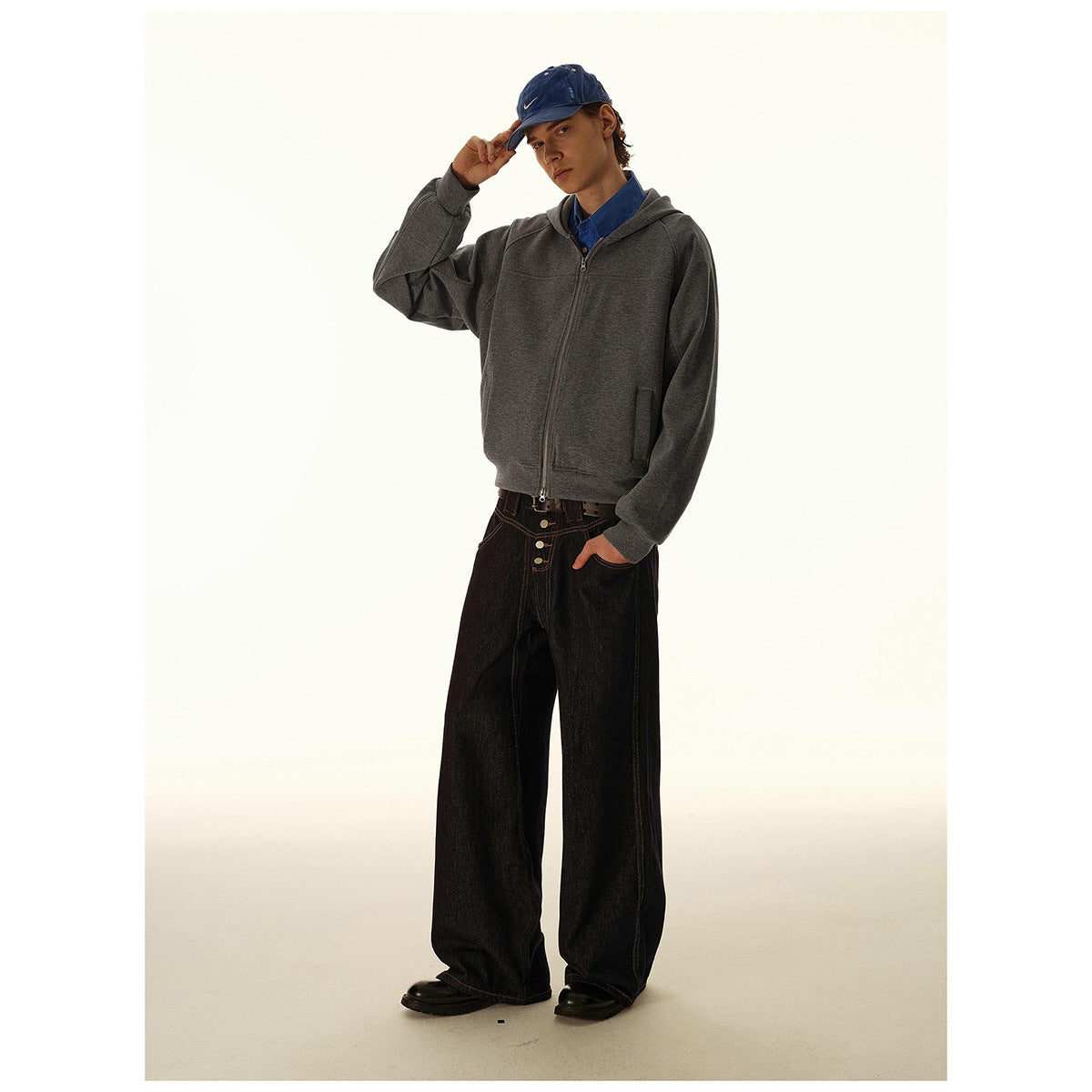 Model wearing a dark gray hooded zip-up jacket with front pockets, shown with dark wide-leg jeans and a blue cap.