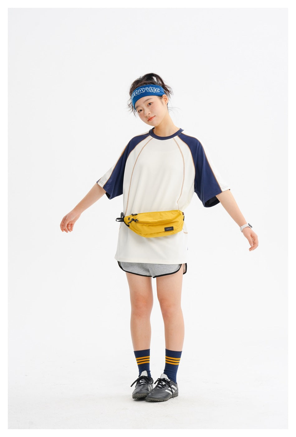 Model wearing an oversized cream athletic tee with navy raglan sleeves and gray shorts, paired with a yellow waist bag.