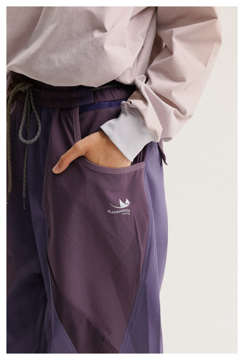 Close-up of purple pants showing the elastic drawstring waist, side pocket, color-block panels, and printed garment logo.