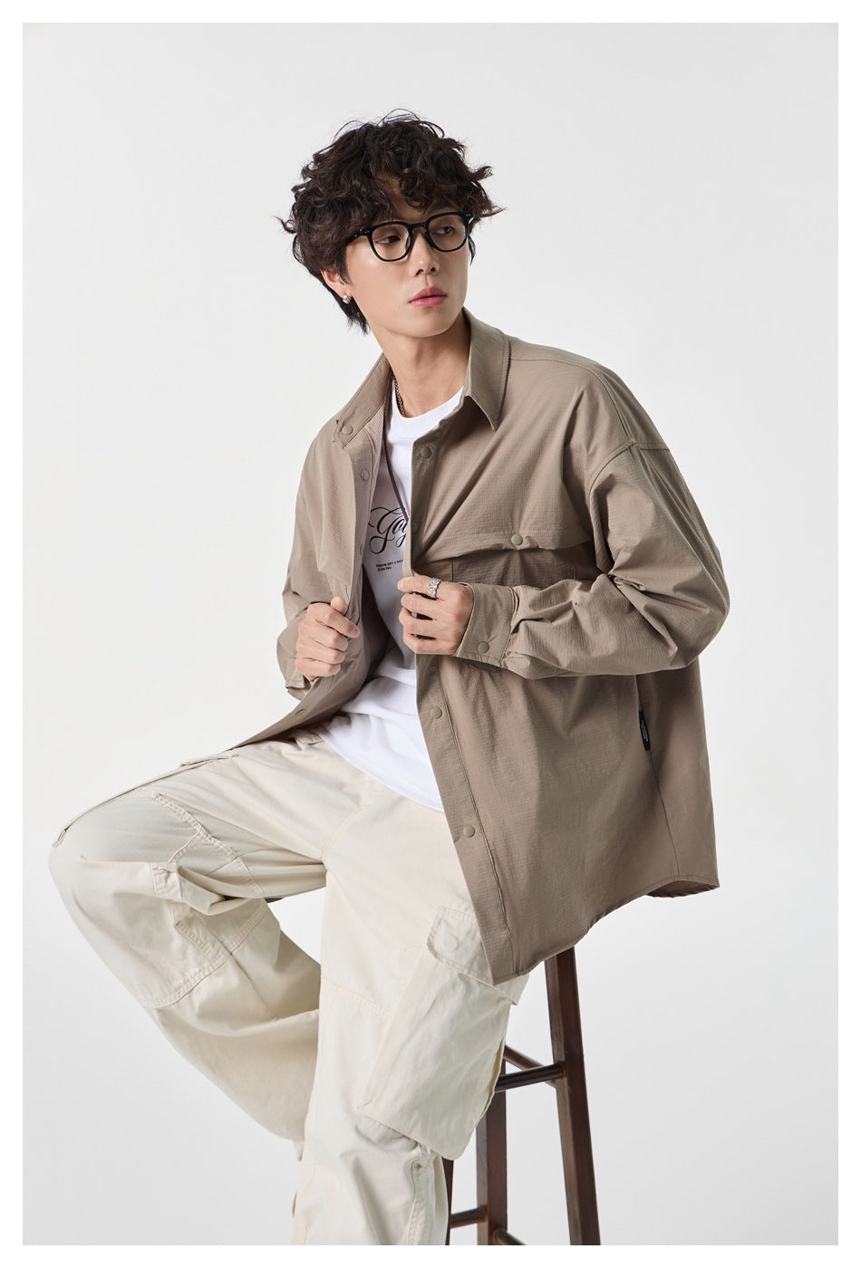 Model seated in a taupe overshirt with snap buttons and flap pockets, white printed tee, and cream cargo pants.