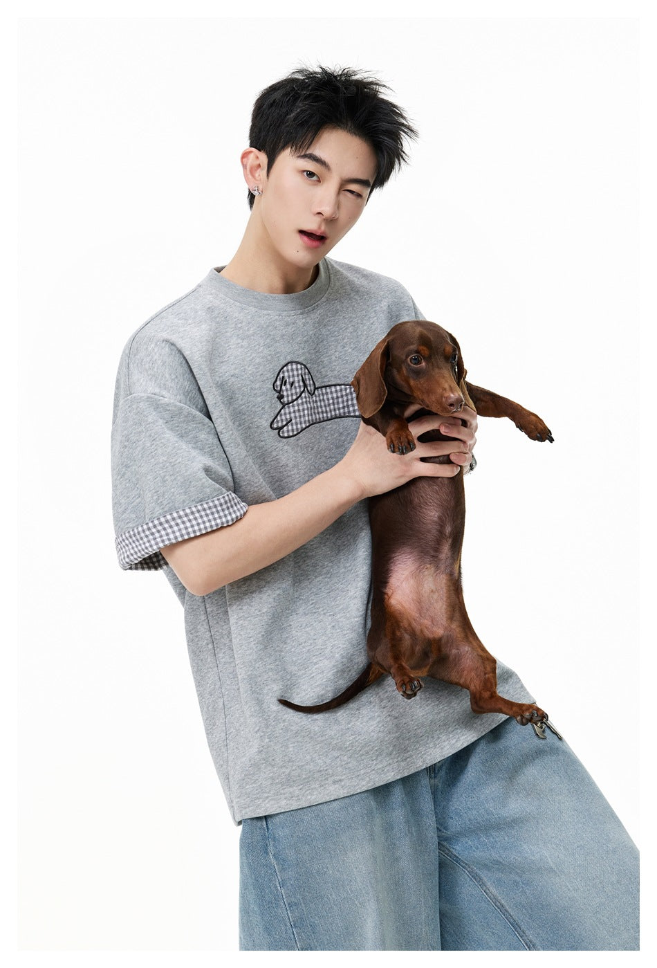 Model holding a brown dachshund, wearing a gray T-shirt with a checked dog graphic and checked sleeve cuffs.