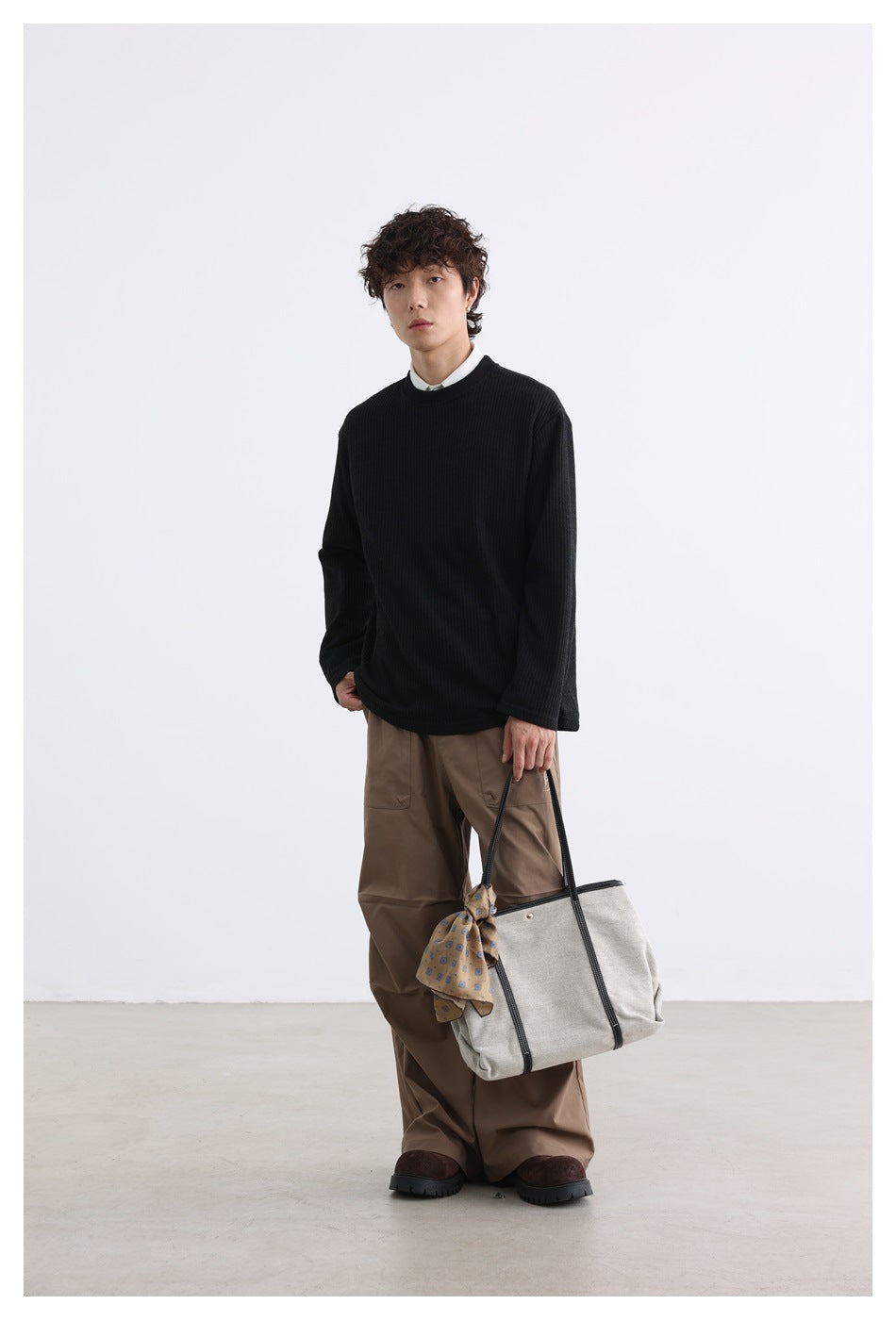 Full-length model wearing a black ribbed crewneck over a white shirt and carrying a light tote bag.