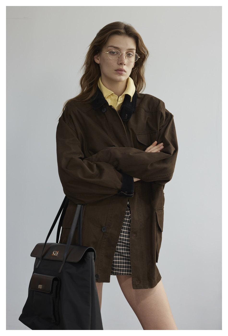 Model wearing an oversized dark brown jacket with a black collar over a yellow shirt and plaid skirt.