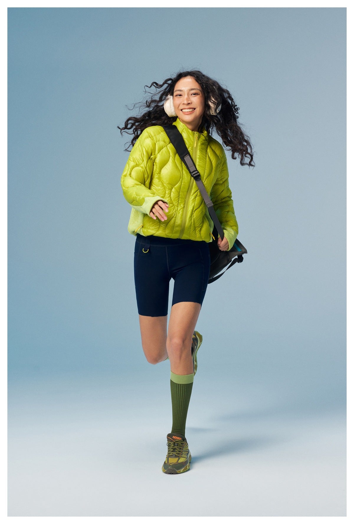 Model running in a yellow-green quilted zip jacket with navy bike shorts and a shoulder bag against a blue backdrop