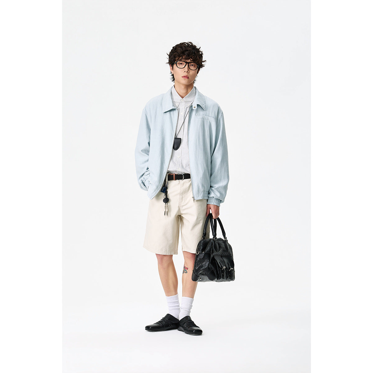 Full-length view of a model wearing a pale blue jacket, checked shirt, cream shorts and carrying a black bag.