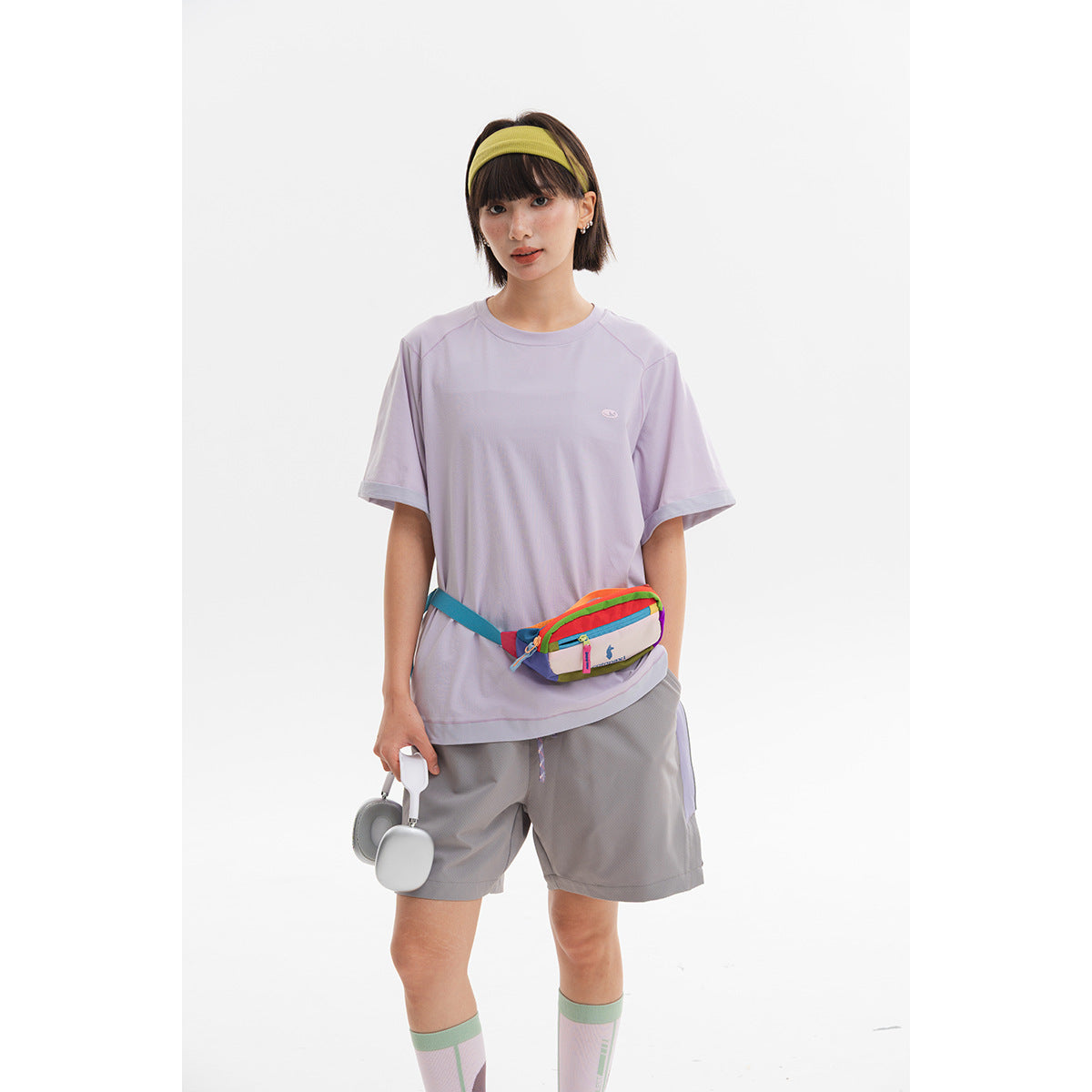 Model wearing a loose pale lavender short-sleeve shirt and gray shorts, with a colorful waist bag.
