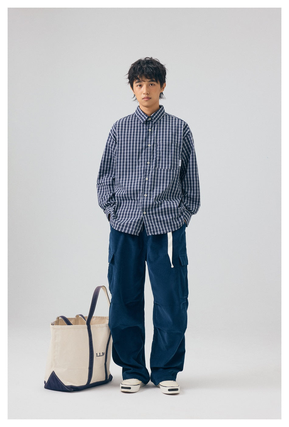 Model standing in deep blue wide-leg cargo pants with a white belt, plaid shirt and sneakers beside a tote bag