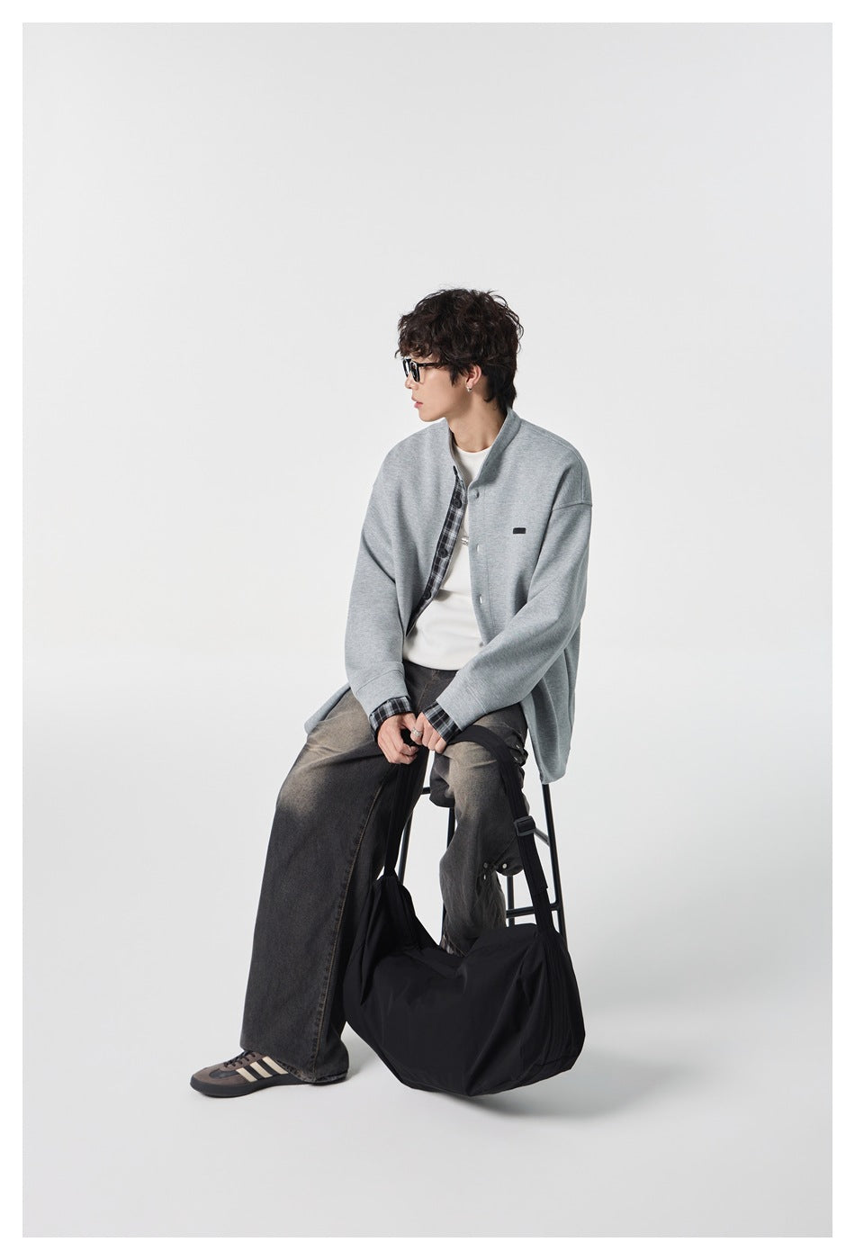 Seated model wearing a light gray jacket with plaid trim and oversized dark jeans; a black bag rests beside him.