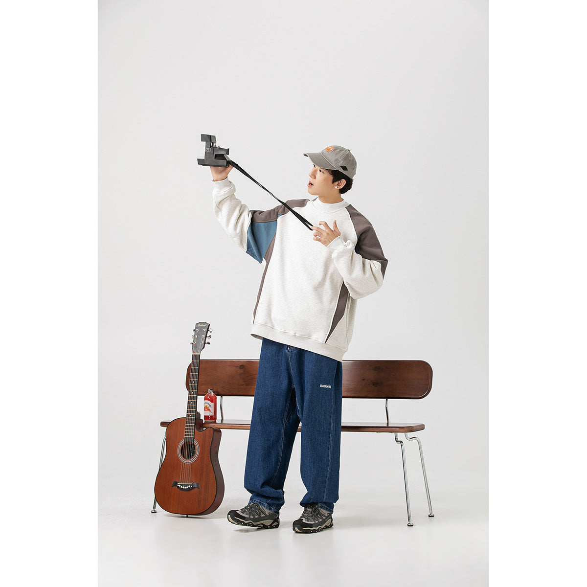 Model wearing an off-white, gray, and blue color-block sweatshirt with loose blue jeans, holding a camera.