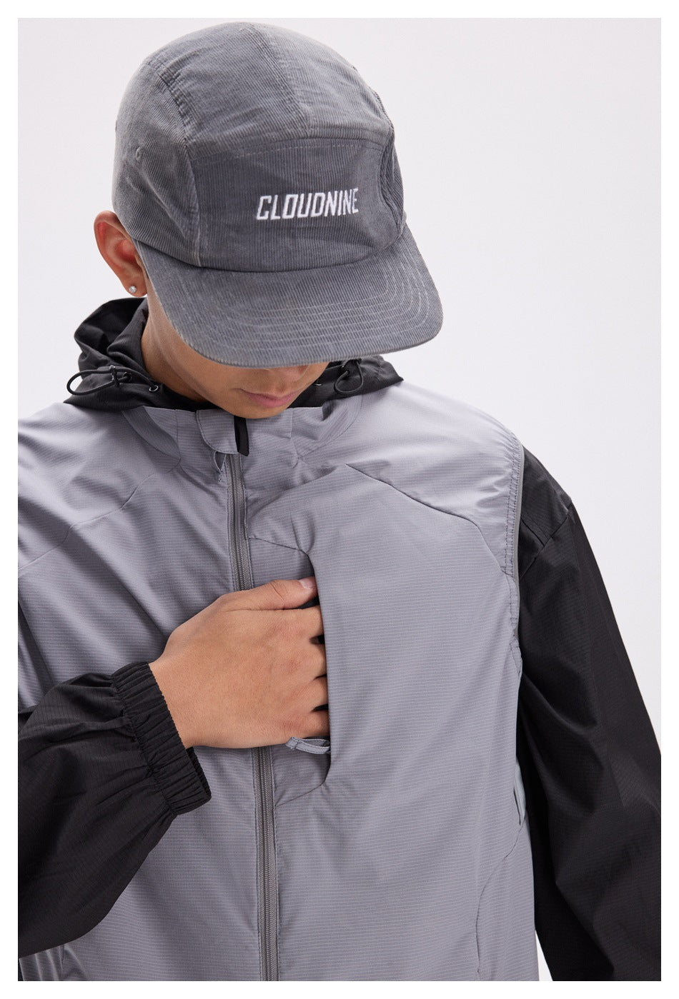 Close-up of a person wearing a gray vest, hand at the diagonal chest pocket, with a gray cap over a black hooded jacket