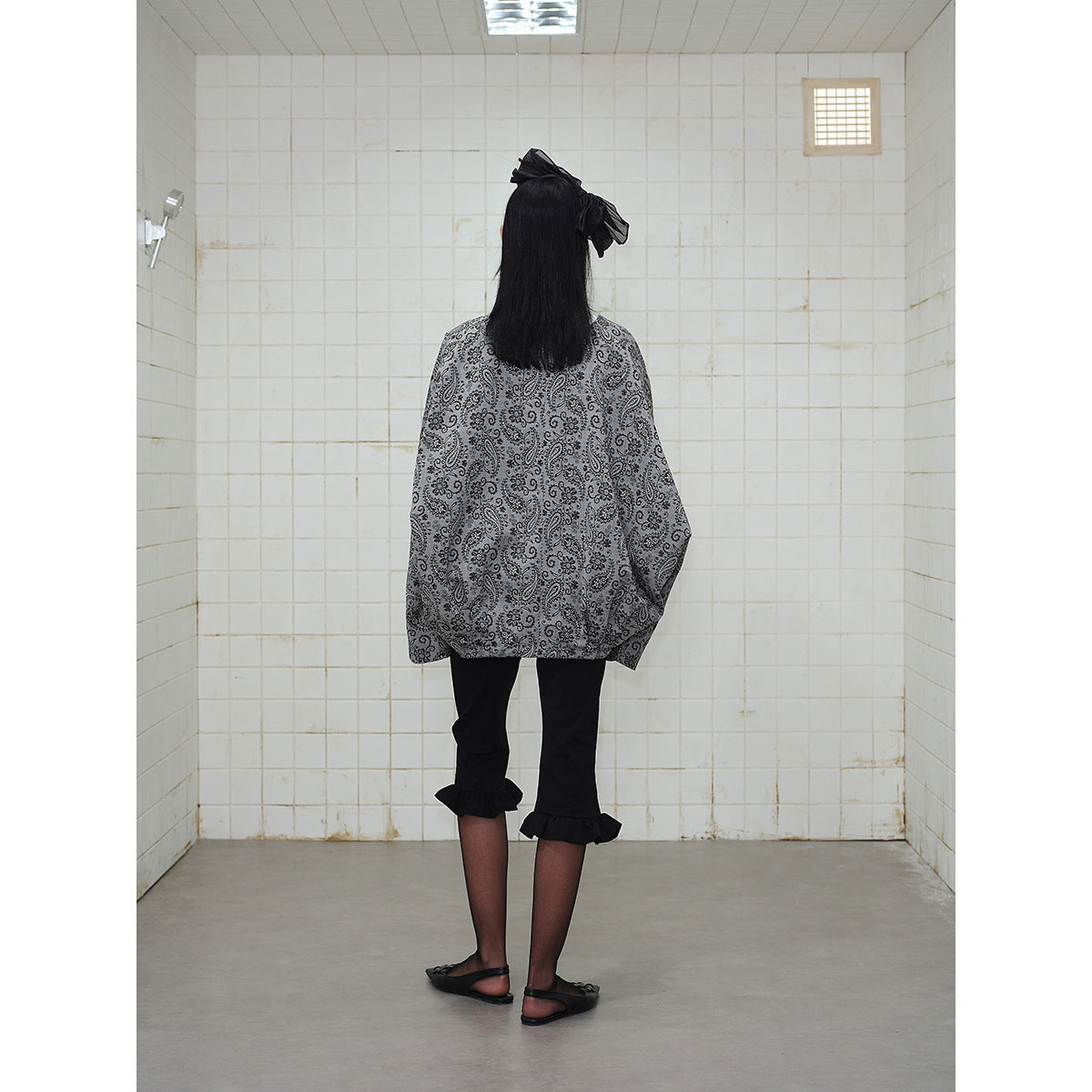 Back view of a model wearing an oversized gray paisley jacket and black cropped pants with ruffled hems.