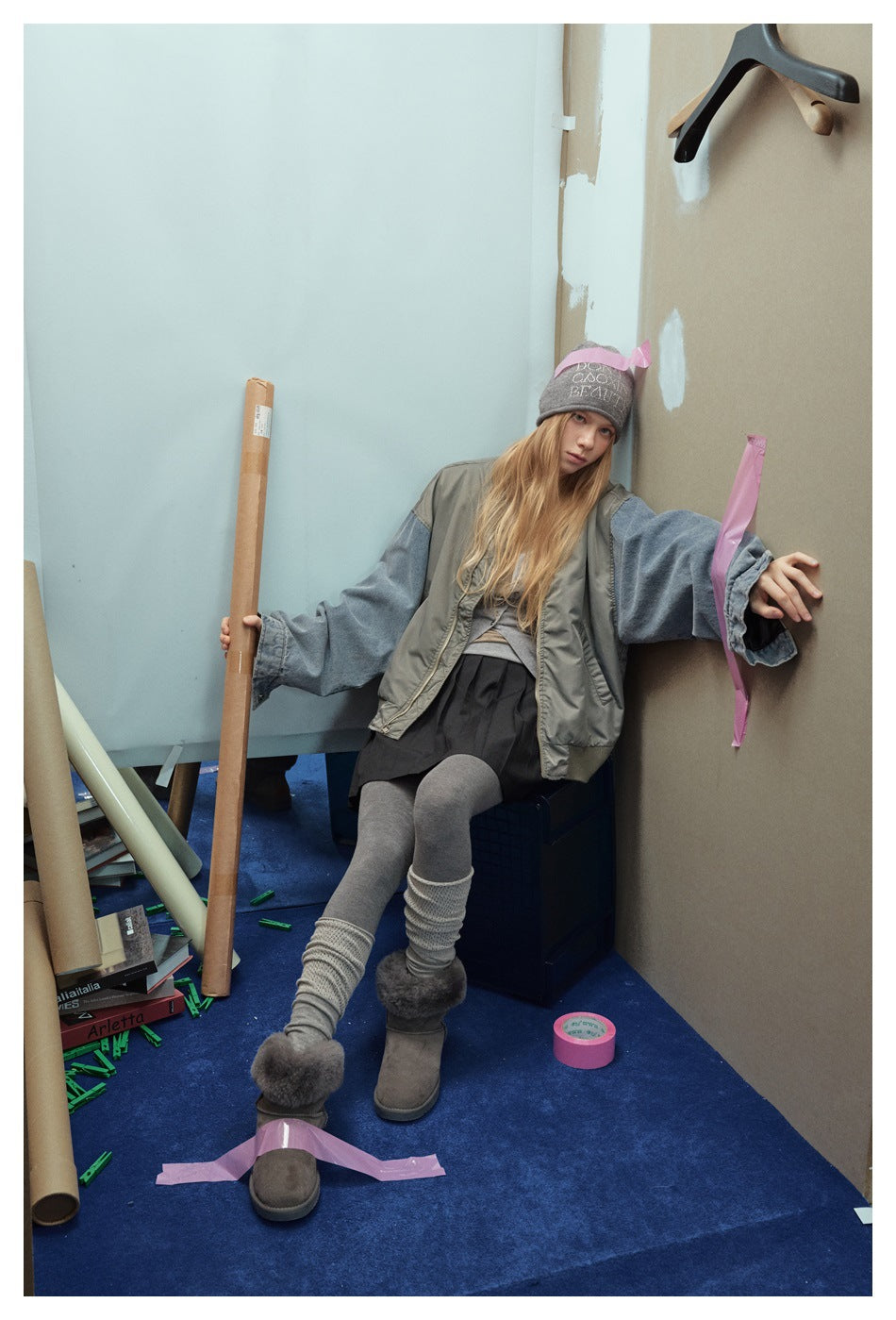 Model seated in an oversized olive-gray jacket with blue denim sleeves, layered over a gray top and black skirt.