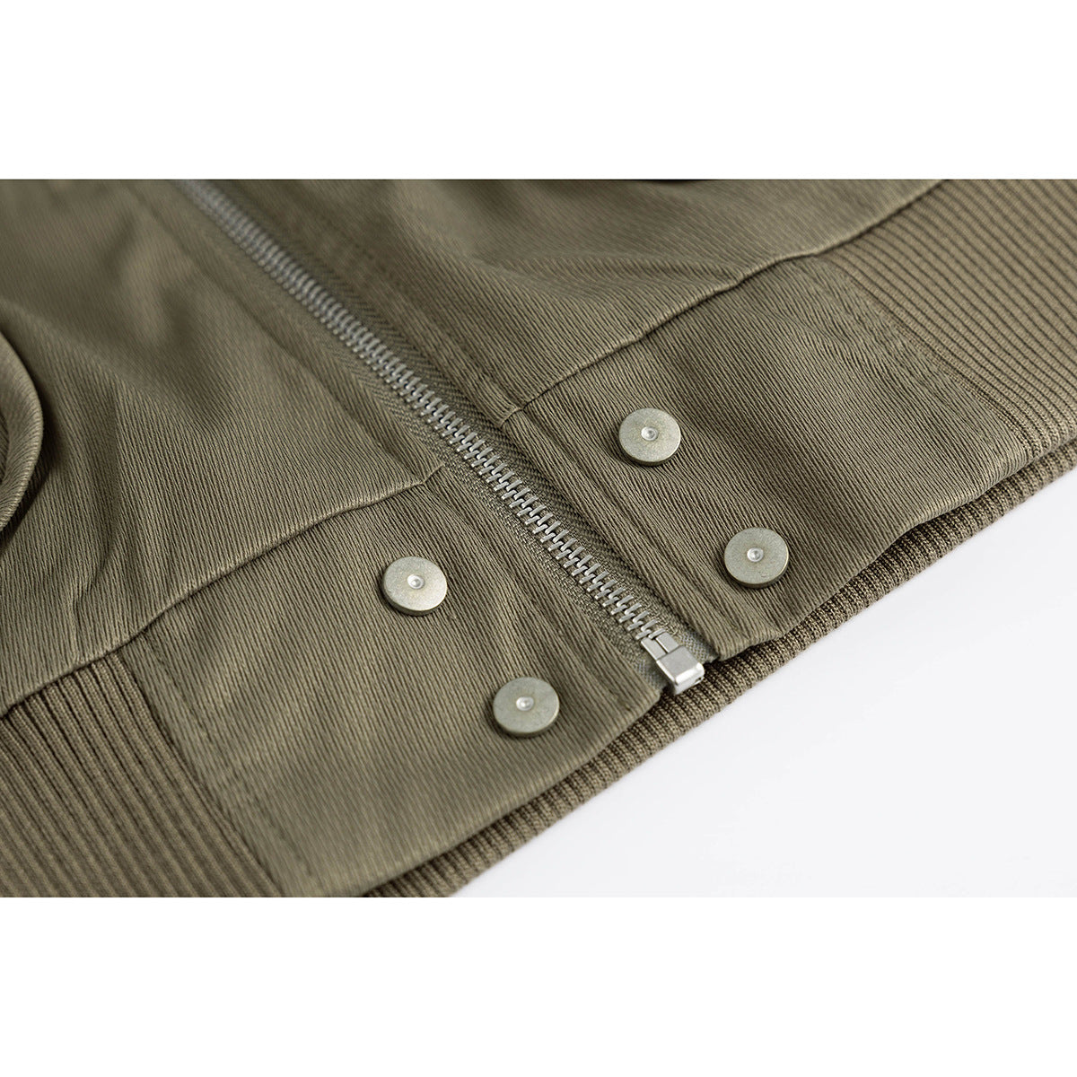 Close-up of an olive jacket’s front zipper, metal snaps, seam and ribbed hem.