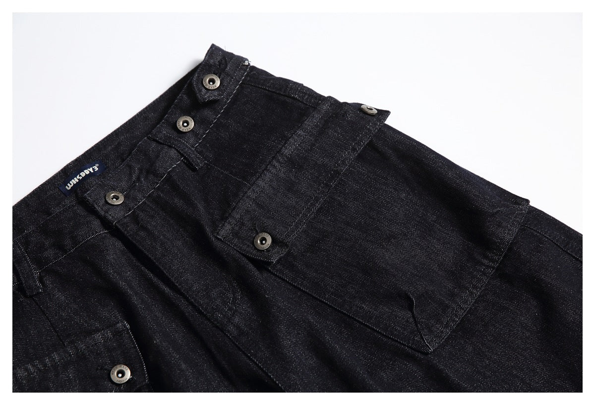 Close-up of the upper front of black denim trousers, highlighting the buttoned waistband and large pocket.