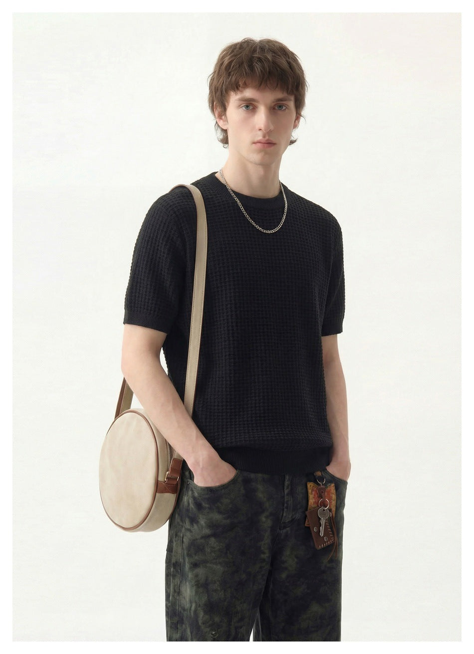Model in a black waffle-knit short-sleeve crew neck top with a chain necklace, hands in pockets, with a round shoulder bag