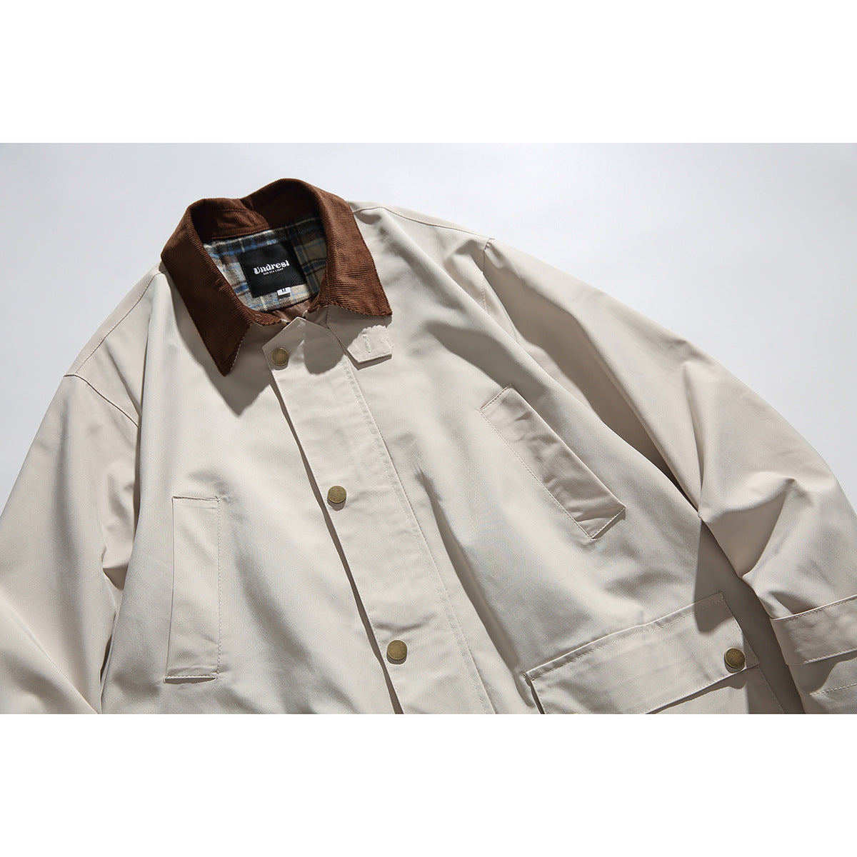 Close-up of a light beige jacket with a brown collar, brass-tone snaps, and large front pockets.