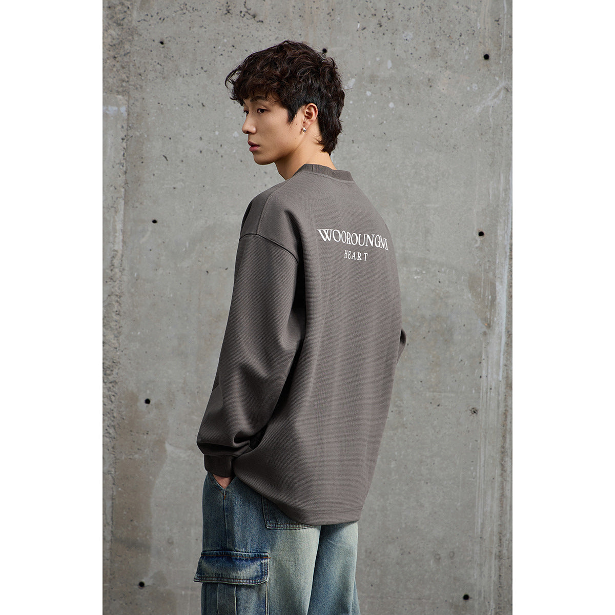 Model shown from behind in a taupe oversized sweatshirt with white back lettering and loose blue jeans.