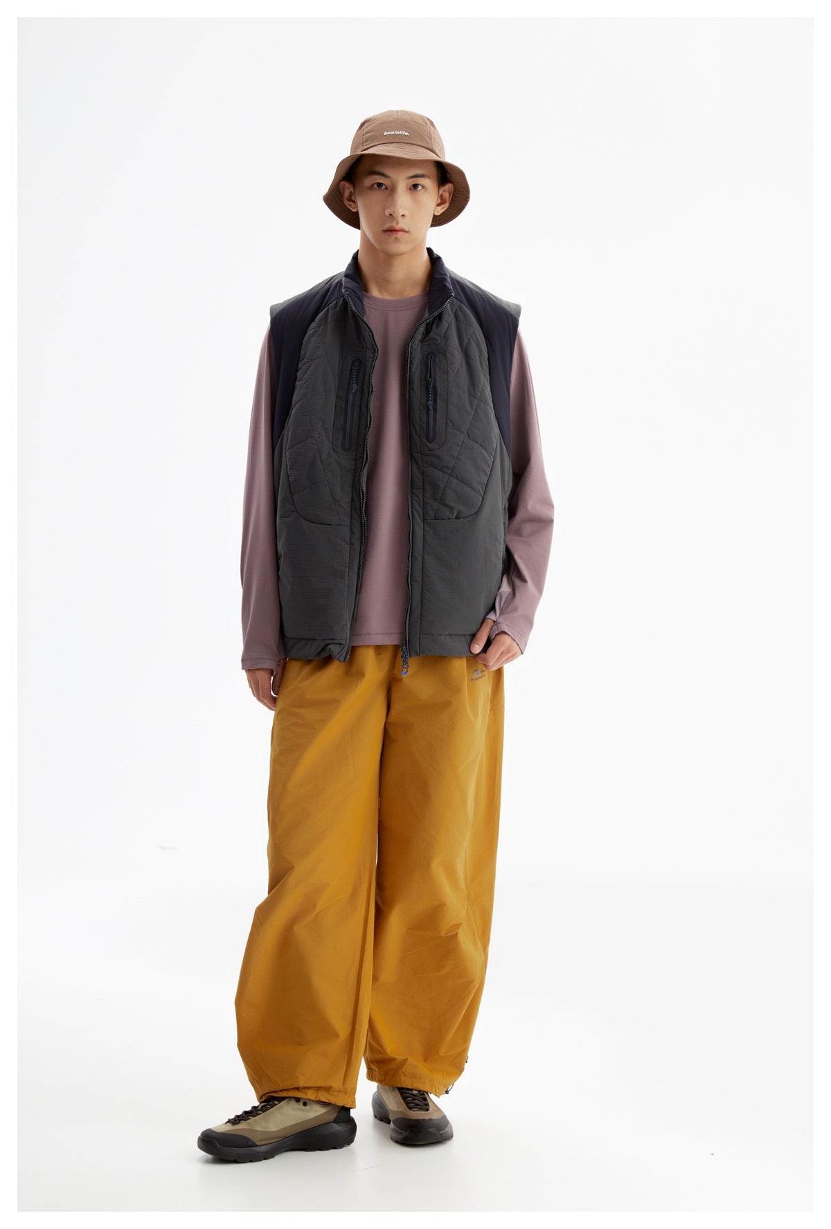 Front-facing model wearing a charcoal quilted utility vest over a mauve shirt and mustard pants.