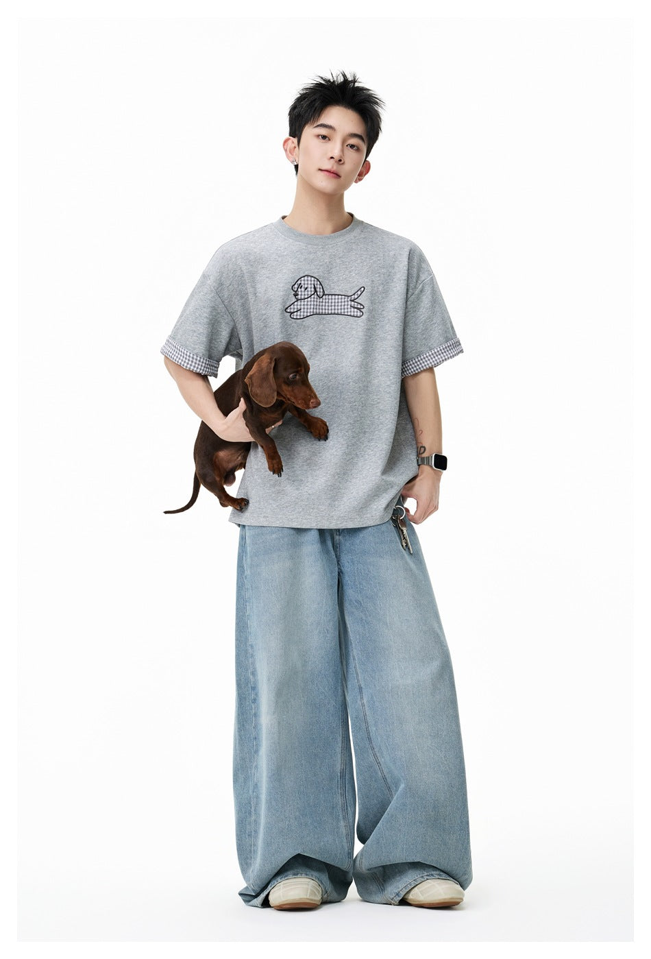 Full-length model wearing a gray dog-graphic T-shirt with checked sleeve cuffs and wide-leg blue jeans, holding a dachshund.