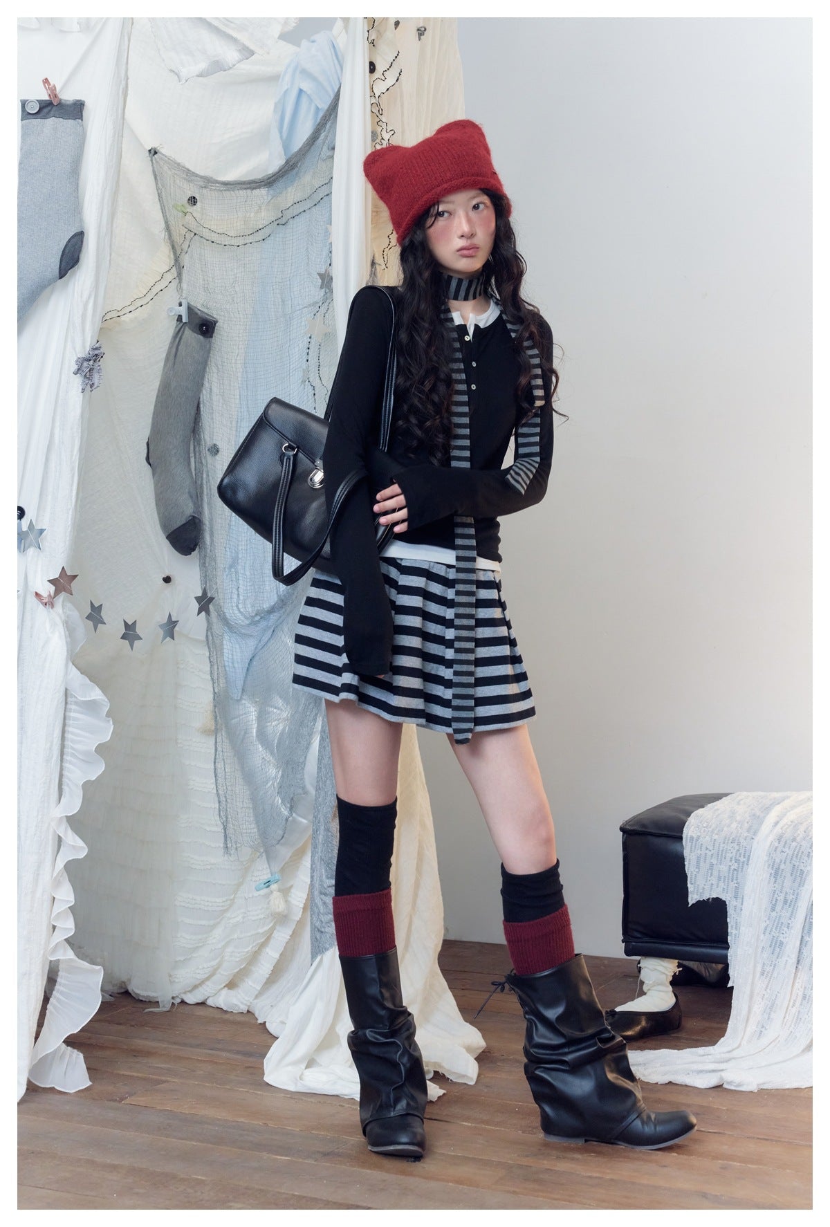 Model poses in a black top, gray-and-black striped mini skirt and scarf, red knit hat, and tall black boots.