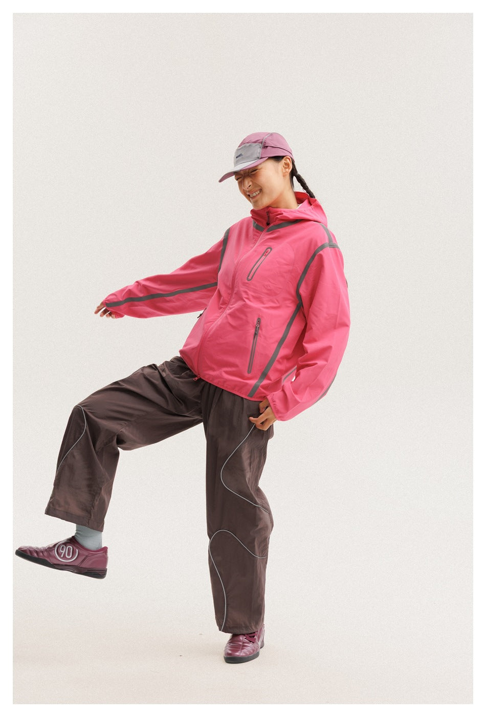 Model wearing a bright pink hooded jacket with gray trim and dark brown trousers, shown from the front at an angle.