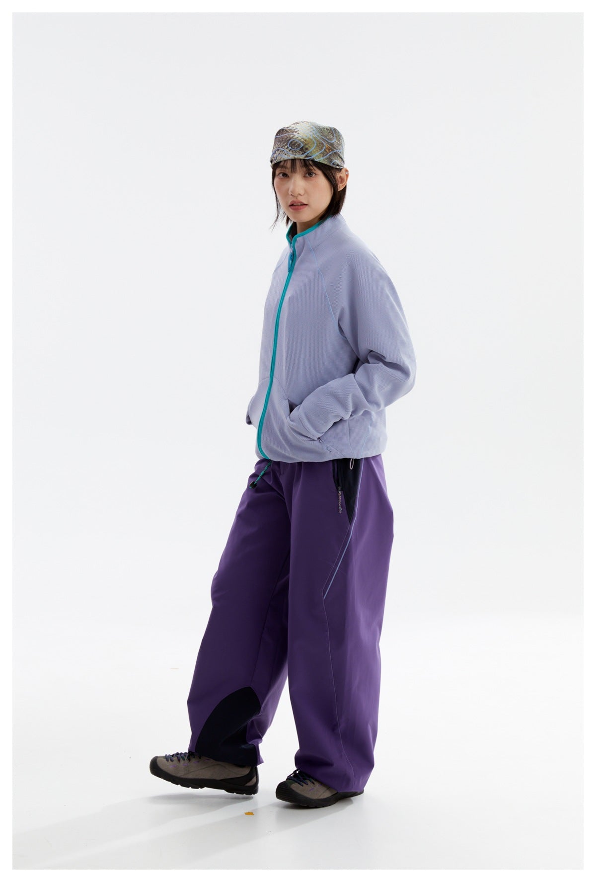 Model wearing a pale lavender zip jacket with teal trim and loose purple pants, shown full length from a three-quarter angle.