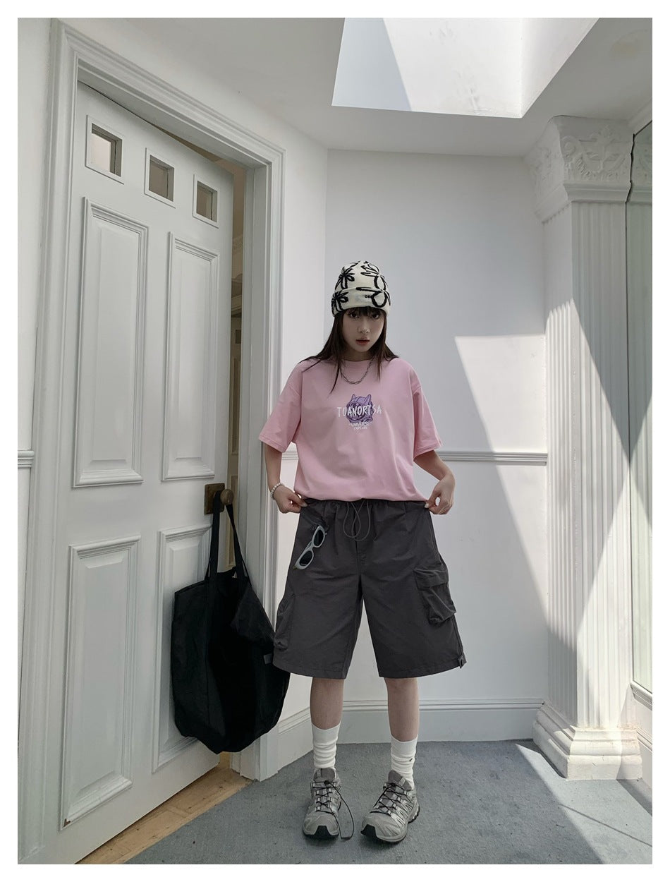 Person wearing dark gray knee-length cargo shorts with a pink t-shirt, beanie and sneakers in a white doorway