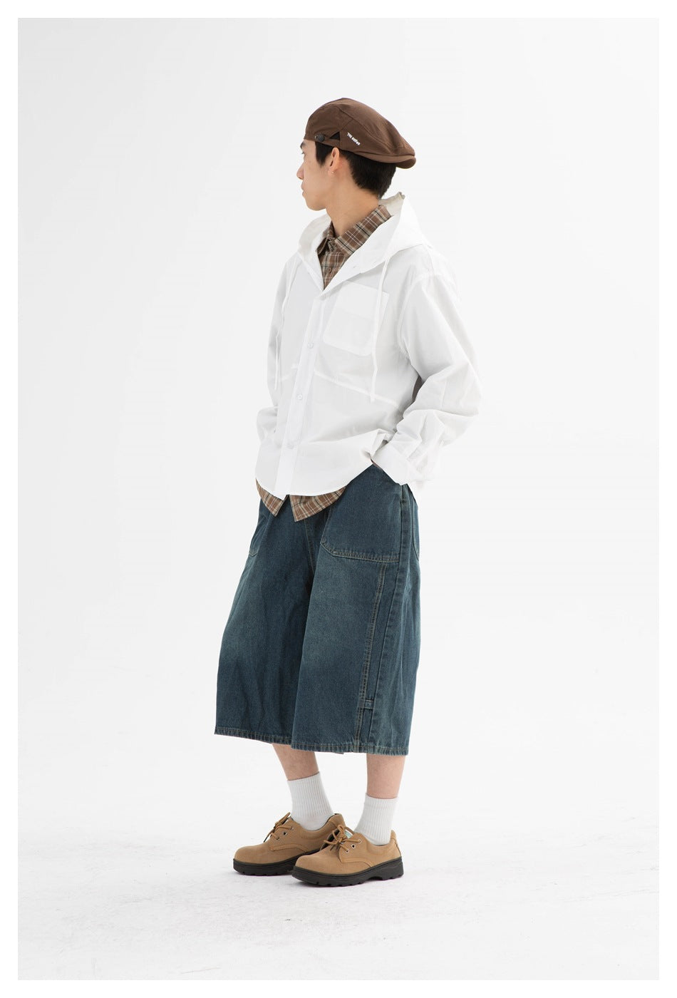 Side view of a model in a white hooded jacket over a brown plaid shirt and wide dark denim shorts.