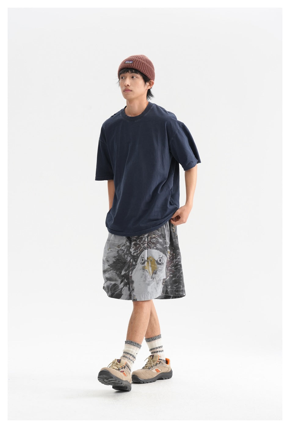 Model in a navy T-shirt and gray eagle-print shorts, shown walking in a rust beanie and hiking shoes.