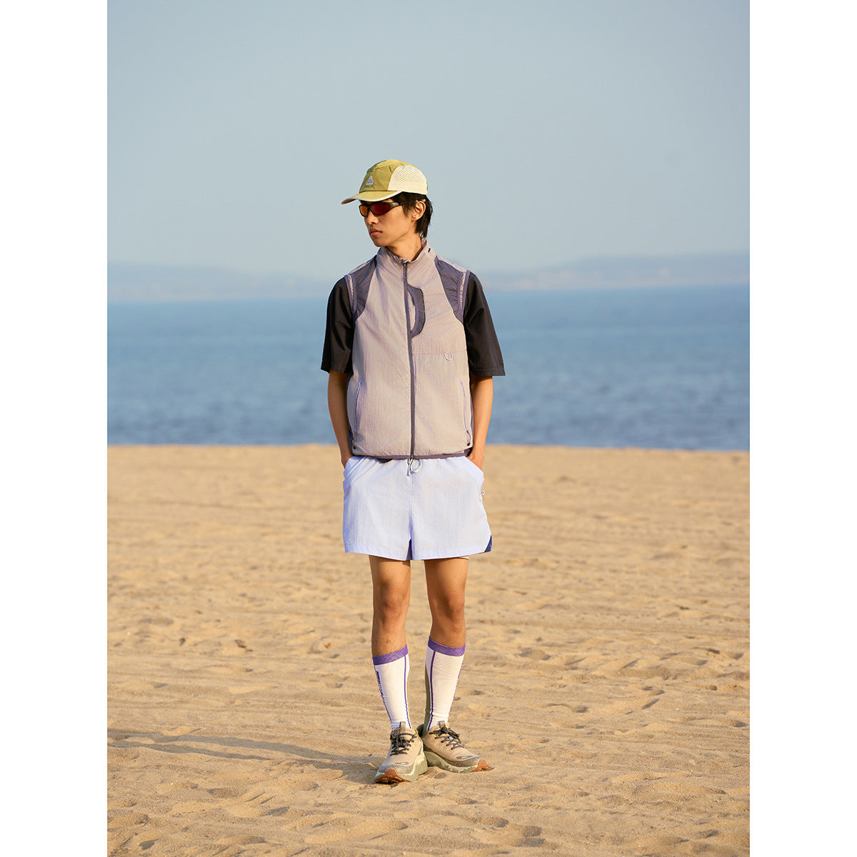 Full-length beach view of a model wearing a light gray-lavender sleeveless zip vest over a black shirt.