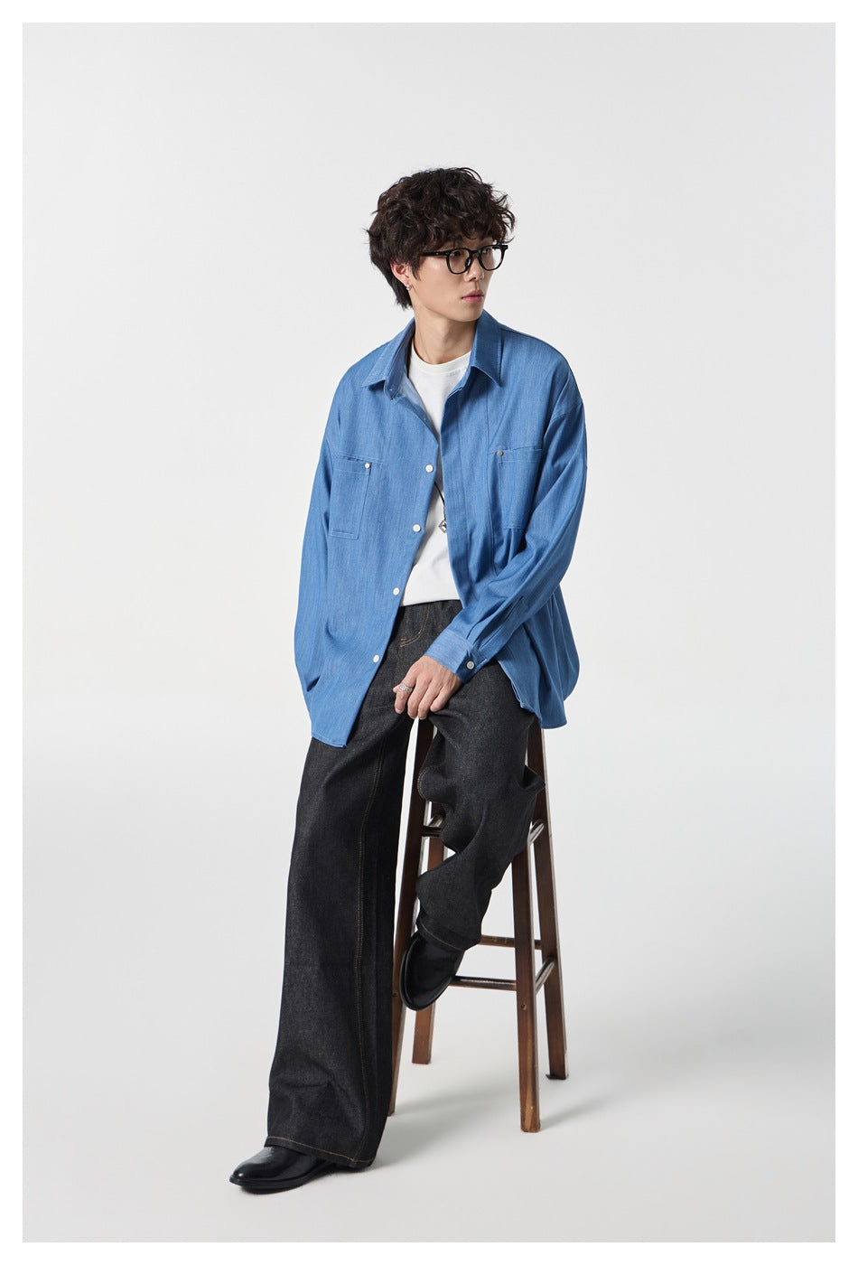 Person seated on a wooden stool wearing an open blue oversized shirt over a white tee with wide dark jeans
