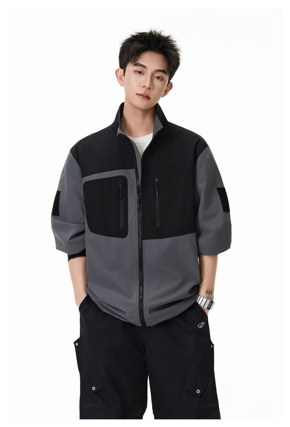 Model wearing a black and charcoal-gray color-block zip jacket with a stand collar and two vertical chest pockets.
