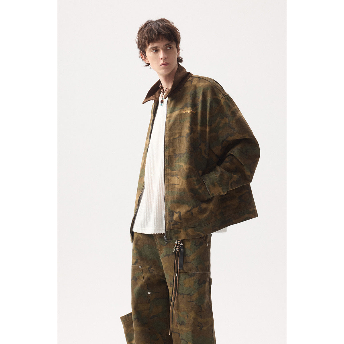 Model in a brown-green camouflage zip jacket with a dark collar and matching loose trousers, shown from a three-quarter