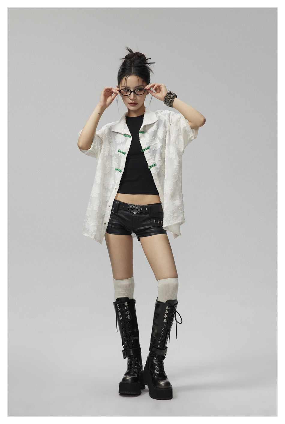 Model wearing an open white floral shirt with green frog closures, styled with black shorts and tall boots.