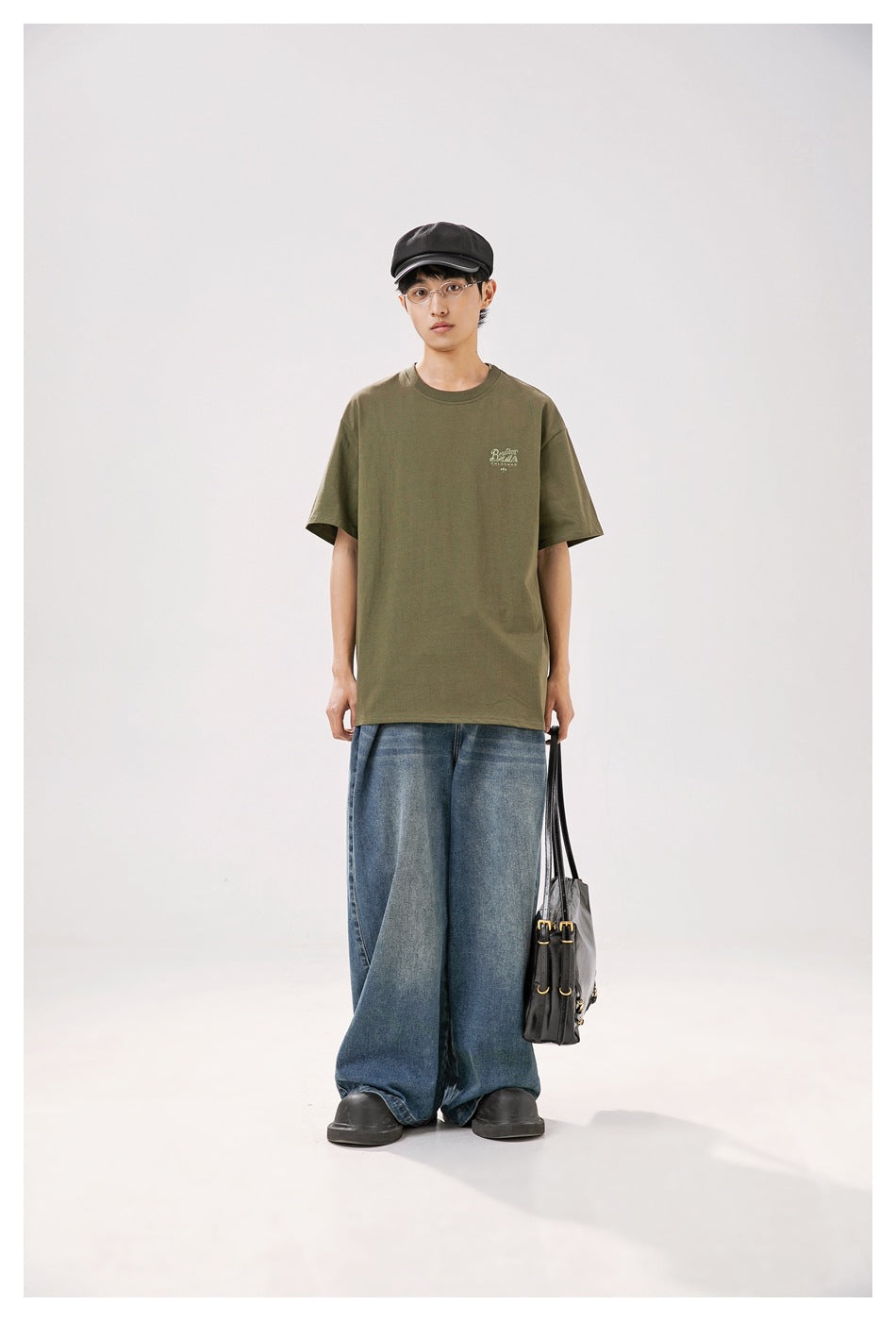 Model wearing an olive-green short-sleeve T-shirt with wide-leg jeans, front view, holding a black bag