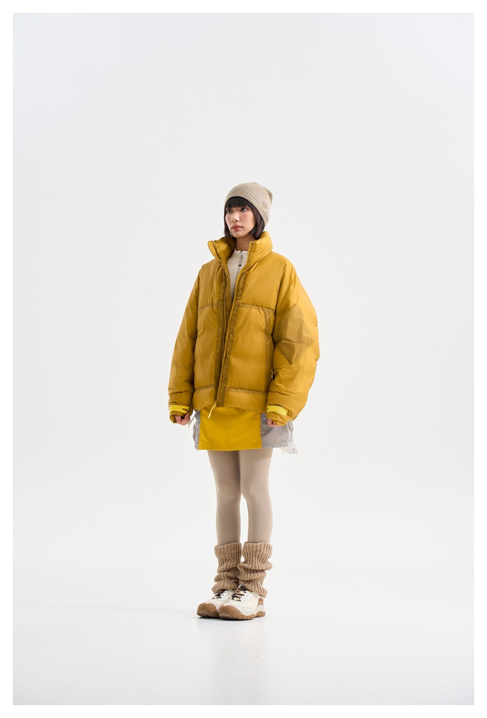 Model wearing a mustard oversized puffer jacket over a yellow tunic, beige beanie, leggings, and slouchy leg warmers.